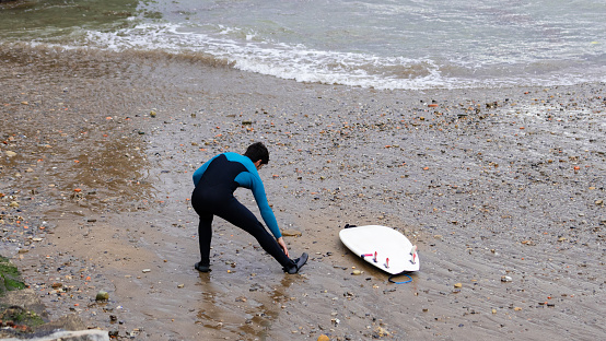 A surfer in a blue wetsuit secures his suit on a pebbly shore before a surf session, with his white surfboard beside him.
