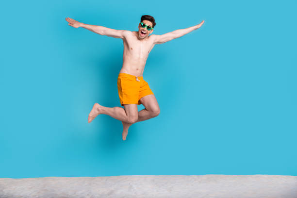 full length photo of nice young male jump fly hands plane dressed stylish yellow shorts isolated on blue color background summer vacation - shirtless men bizarre male imagens e fotografias de stock