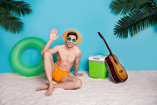 Full length photo of friendly positive guy swimwear sunhat sitting beach waving arm hello isolated blue color background.