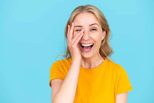 Smiling woman covering one eye with hand and peeking another at camera. Isolated on blue background. Positive emotions concept. Happy, good mood in comic situation.