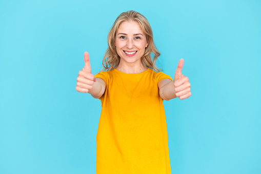 Smiling pretty young woman showing thumbs up gesture isolated on blue background. Happy positive female with like sign recommend service or product. Good choice