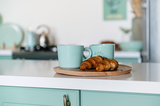 French fresh bakery and coffee on breakfast. Closeup of sweet croissants and cups with drink on wooden tray on white countertop of cooking island on blurred modern kitchen background