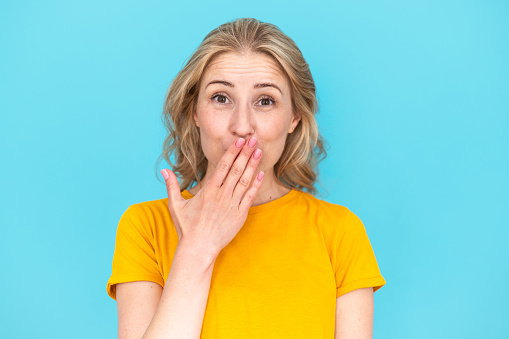 Embarrassed situation, trouble, funny moment, human emotions. Portrait of shy smiling woman covering her mouth with hand, make mistake, look at camera standing isolated on blue copy space background