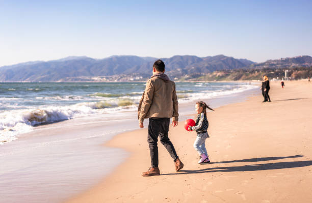A young girl and her Dad playing together at the beach with a ball.