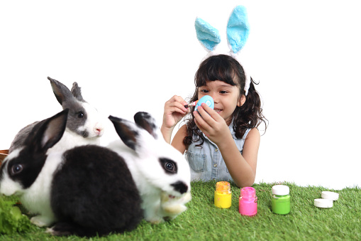 Cute little girl with blue bunny ears painting Easter eggs with brush on green grass meadow, joyful kid being surrounded by her rabbit pet celebrating Easter holiday on white background. Happy child