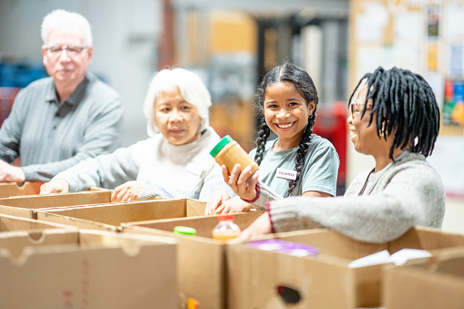 A small group of Volunteers are seen working together at a local food bank as they pack donations.  They are each dressed casually and smiling as they work diligently together.