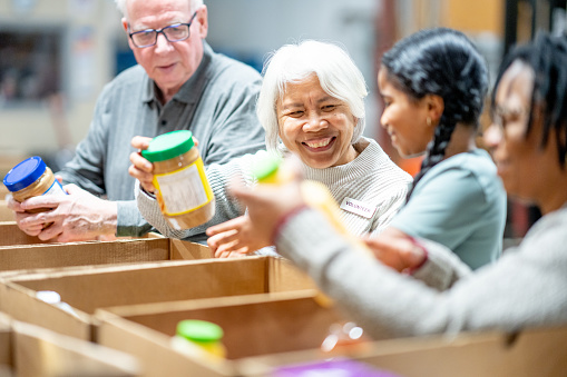 A small group of Volunteers are seen working together at a local food bank as they pack donations.  They are each dressed casually and smiling as they work diligently together.