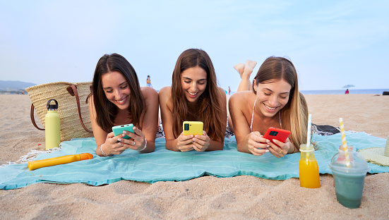 Three caucasian women friends lying on a beach towel during summer vacations in the Caribbean, using smart phone gossiping, online texting and checking social media, laughing and having fun.