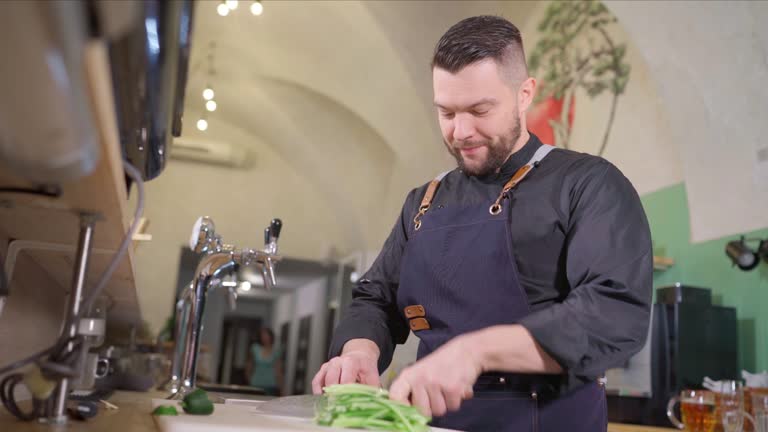 Smiling bearded chef cook dressed black uniform with an apron chopiing a fresh green cucumber with sharp knife. Professional occupation, hard work, food preparation and restaurants industry 4K footage