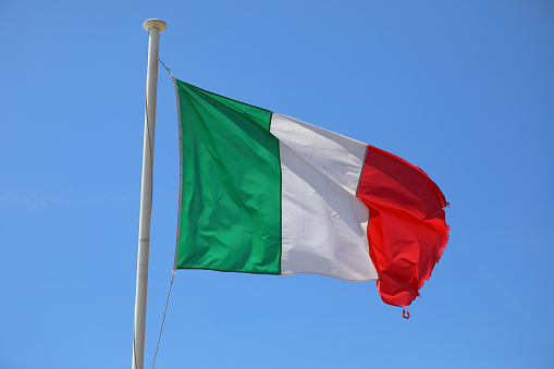 The flag of Italy waving in the breeze on a flagpole against a blue sky on a sunny day.