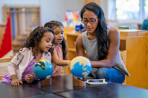 A Montessori teacher sits with two young children as she teaches them about different places in the World using a globe.  The little girls are dressed casually and appear inquisitive.
