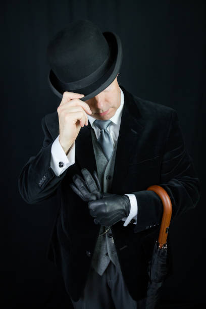 Man in a formal suit, tie, sunglasses, bowler hat, and open umbrella sits on a chair, on a country road
