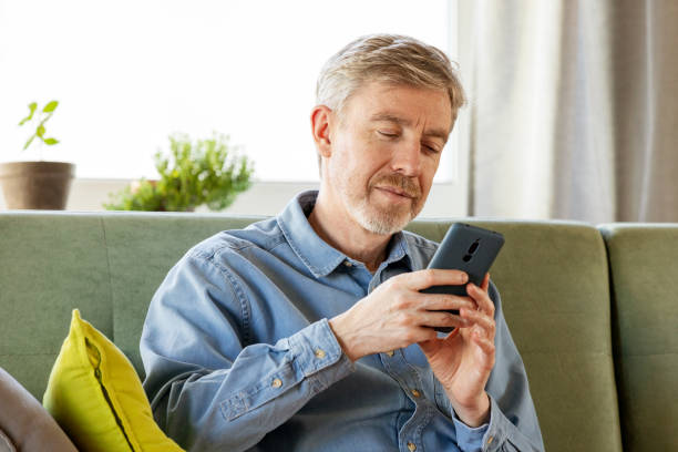 istock 50 year old man sitting on the sofa in his home using a smart phone 2051200033