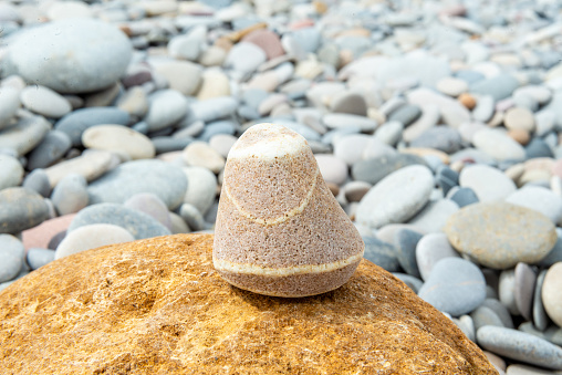 A striped cone-shaped stone stands atop a sandy rock, surrounded by grey pebbles