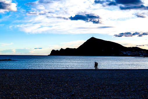 Altea, Alicante - Spain - 01-05-2024: Two silhouettes stroll on a pebble beach in Altea with a mountain backdrop under the evening sky