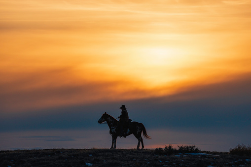 Horseback riding on a Wyoming horse ranch in the winter on a rural landscape, as the sun sets and casts the rider in silhouette.