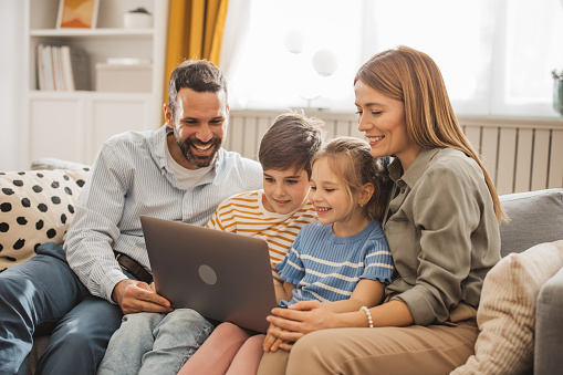 Mother and father using  laptop with their children on the sofa at home. Online shopping or talking on video call.