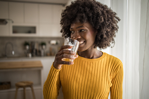 Portrait of a smiling young beautiful black woman with an afro hairstyle holding a glass of fresh clean drinking water. Concept of healthy lifestyle and fluid intake