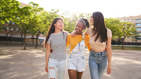 Three cheerful stylish and friendly multi-ethnic girls walking embracing city park and laughing together. Happy group of attractive women having fun outdoors. Generation z people community concept