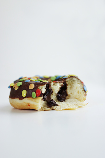 One bitten Donut glazed with chocolate and colorful smarties isolated on white background. Pieces of doughnut with colorful chocolate nibble on top. Side view. Copy space.