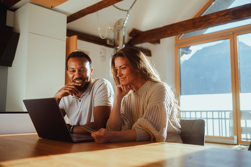 A couple is sitting together at a wooden table in their living room, engaged in a collaborative task on a laptop, with the warm sunlight and a view of the mountains through the window creating a pleasant backdrop.