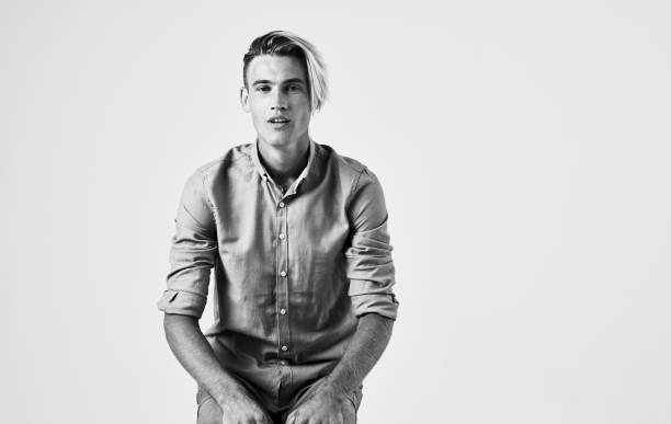 Black and white portrait of a young handsome man in a white shirt, black tie and vest, sitting and looking at the camera, against plain studio background
