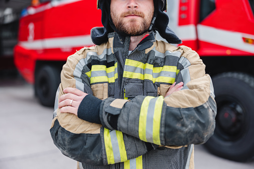 A Caucasian firefighter in his late 20s standing in front of a fire truck, wearing a fire protection suit and helmet.