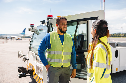 Diverse Hispanic male and Asian female airport workers talking in front of a push back tug at the airport runway.