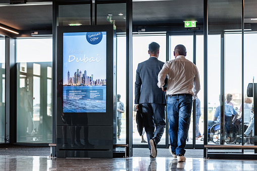 Full length shot from behind with blurred background of two men walking and about to cross the exit door of the airport, the one on the right is a Latin-American driver who arrived to pick the other one up.
