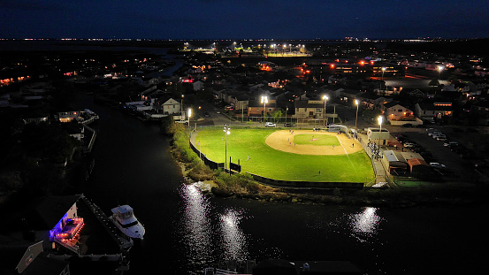 Baseball sports field in light on waterfront of a small town with houses and boats at night. Sports and leisure activity on sea banks: drone night view