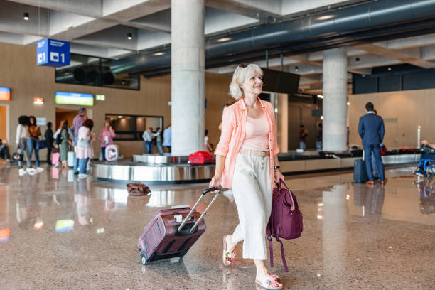 Blonde Senior Woman Pulling Her Suitcases Through The Airport Full length shot with blurred background of a smiley blonde senior woman pulling her belongings through the airport while looking for the exit door to meet her daughter who is waiting for her. walking into glass door stock pictures, royalty-free photos & images