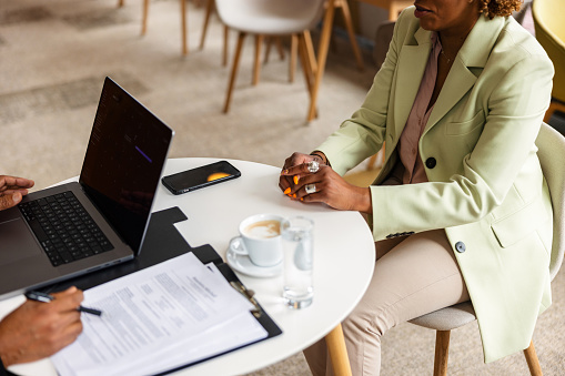 Unrecognizable black mature businesswoman sitting in a cafe, being interviewed by a black person with a laptop and notes. The interviewer is taking notes while she is speaking. The woman is dressed in businesswear.