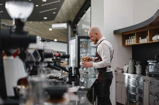 Side view of an experienced adult male barista making a coffee with a pour-over technique. The shot was taken behind the counter. The man is surrounded by various coffee-making machines and glassware. The barista look focused and professional.