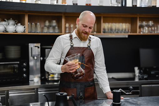 An adult Caucasian male barista holding a glass with a coffee filter in it while the coffee is dripping through the filter. The barista looks relaxed and patient. He is wearing a brown leather apron and a professional-looking shirt. The man is located by the steel counter surrounded by glassware.