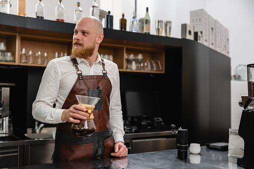 Adult Caucasian male Barista standing by the steel counter and holding a glass full of pour-over coffee he has just prepared. The man is wearing a leather apron. He is looking away from the camera.