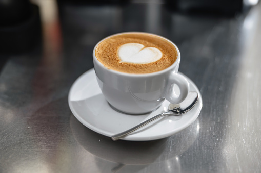 A charming cup of cappuccino coffee with heart-shaped latte art standing on a steel countertop. The coffee cup is minimalistic and white.