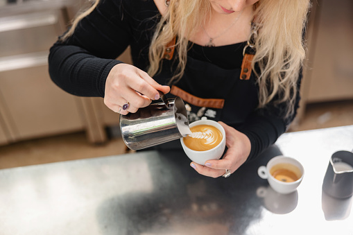 A Caucasian female barista with long blonde hair making coffee for a customer at a specialty coffee shop. She is making a heart-shaped latte art. The cup of coffee is in the focus. The barista's face is not visible.