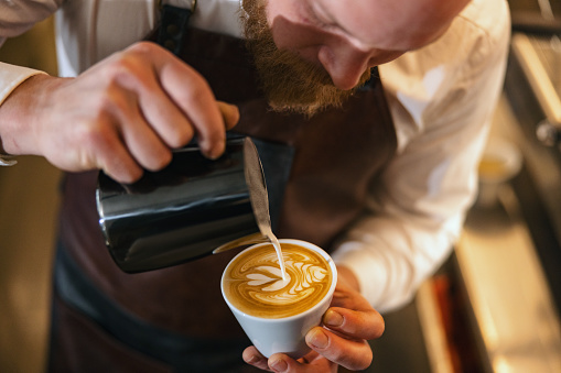 Close up of a Caucasian man's hands holding a cup of coffee and pouring in the frothed milk in order to create beautiful latte art. The man is precise and patient while making the coffee for the client. He is wearing an apron to protect his clothes.