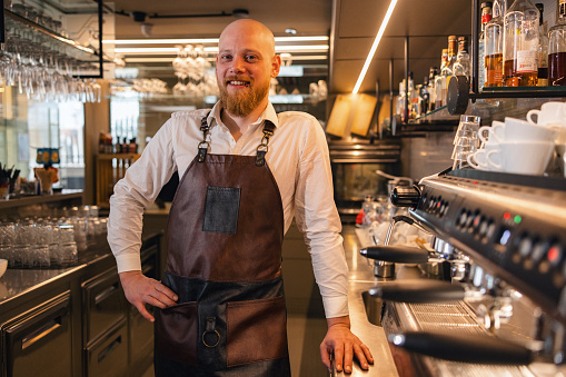 A cheerful adult Caucasian male barista wearing an apron standing by the counter and smiling at the camera. He looks happy and joyful. He is surrounded by coffee-making appliances and dishes.