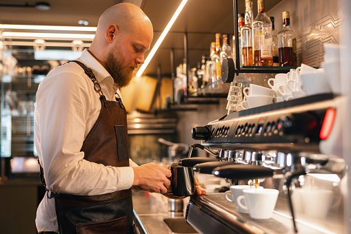 A professional adult male barista using the coffee maker at a cafe to prepare two cups of coffee for his customers. He is wearing a brown apron while working to protect his clothes. Due to the hot temperatures, there is steam as the coffee machine is running.