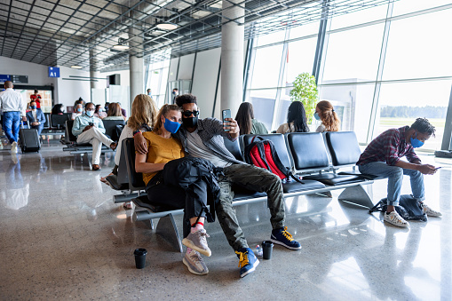 A carefree and cheerful couple, dressed casually and wearing face masks, sitting in an airport waiting area, taking a fun selfie together. The man, in sunglasses, and the woman are digital nomads, excited for their upcoming adventures.