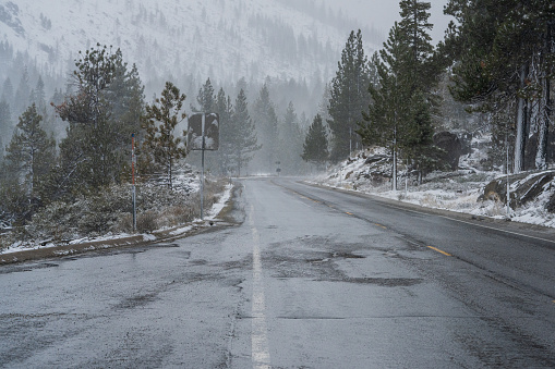 Looking downhill at wet mountain road with forest backdrop and snow during winter storm.