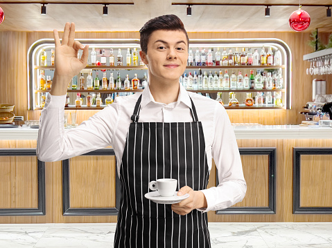 Young waiter holding an espresso coffee cup and gesturing ok sign at a bar