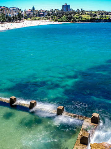 Sydney, Australia - Nov 28, 2022: Powerful white waves (blurred motion) crashing into the Ross Jones Rockpool at Coogee Beach. Ocean water showing tropical blue and green colors of the shallow floor.