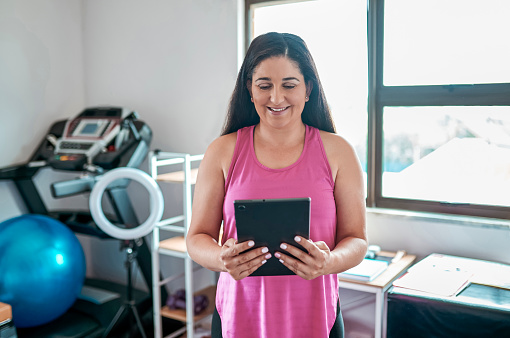 Mature mother with long hair and wearing pink tank top watching and learning from online workout tutorial on digital tablet and smiling happily in home gym
