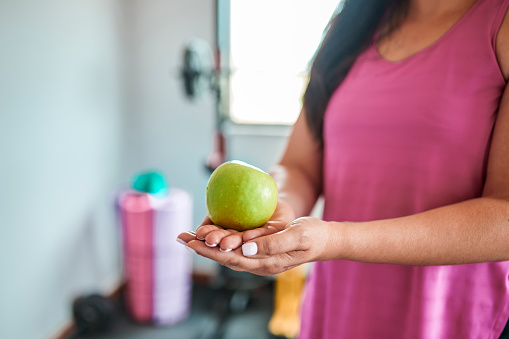 Woman with long hair and wearing pink tank top holding out green apple on cupped hands, standing in home gym