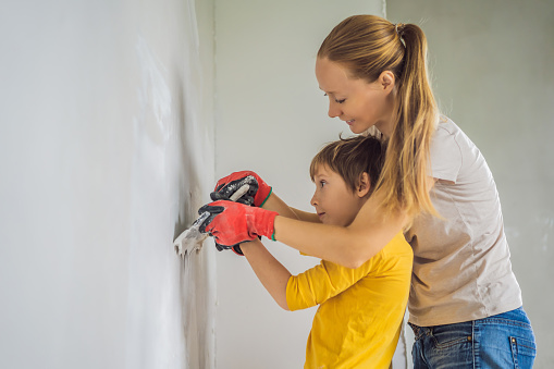 Woman with hir son makes repairs at home, she teaches boy to plaster the walls with a spatula in his hands.