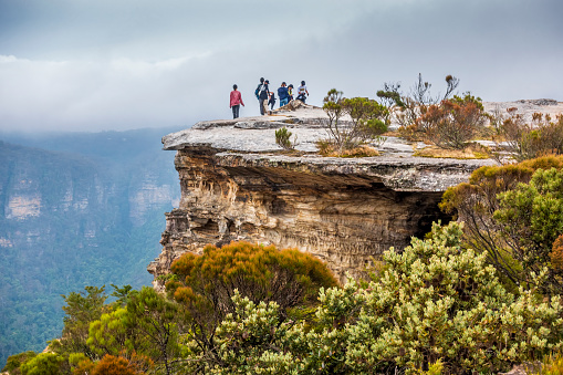 Sydney, Australia - Dec 29, 2022: Rugged rocky cliff with visitors on the plateau. Australian heath or shrubland habitat in Sydneys Blue Mountains National Park. Low lying fog in the distance.