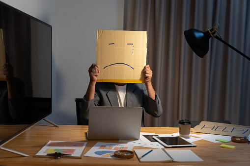 Portrait of businessman covering his face with paper mask with sad face drawn on it, bad negative sullen depression anonymouse person.