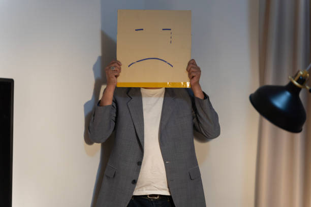 employé malheureux avec une boîte à la place de sa tête. portrait d’homme d’affaires couvrant son visage avec un masque en papier avec un visage triste dessiné dessus. - sadness human face depression smiley face photos et images de collection